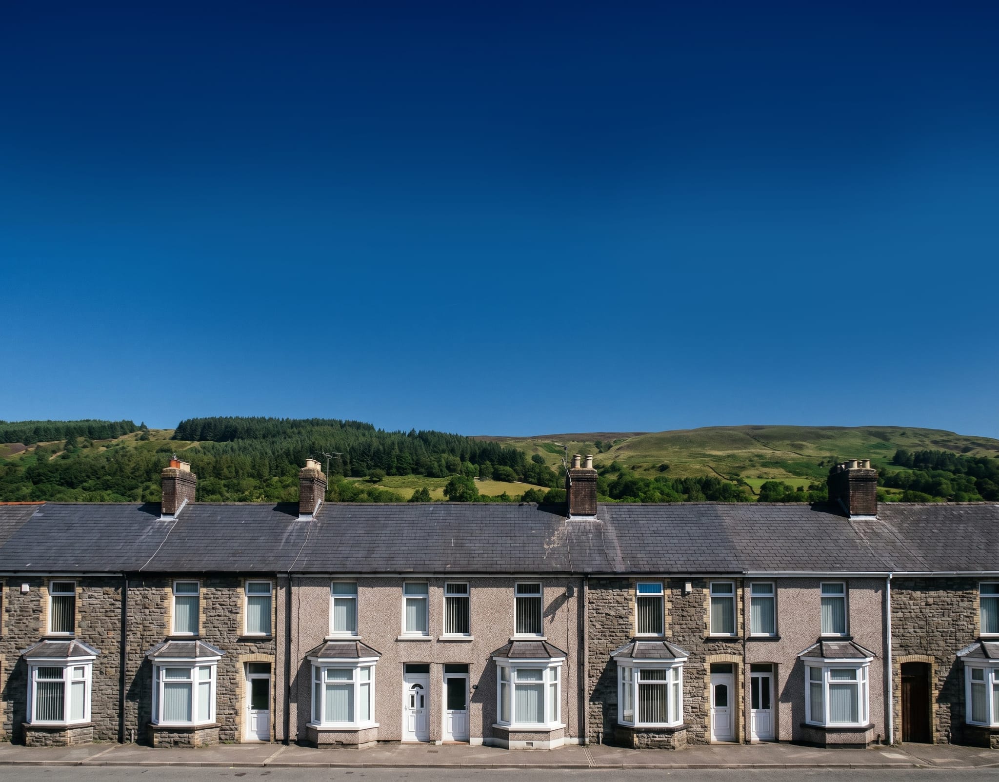 A row of stone terraced houses with slate roofs and chimney stacks, below a green hillside in the South Wales valleys.